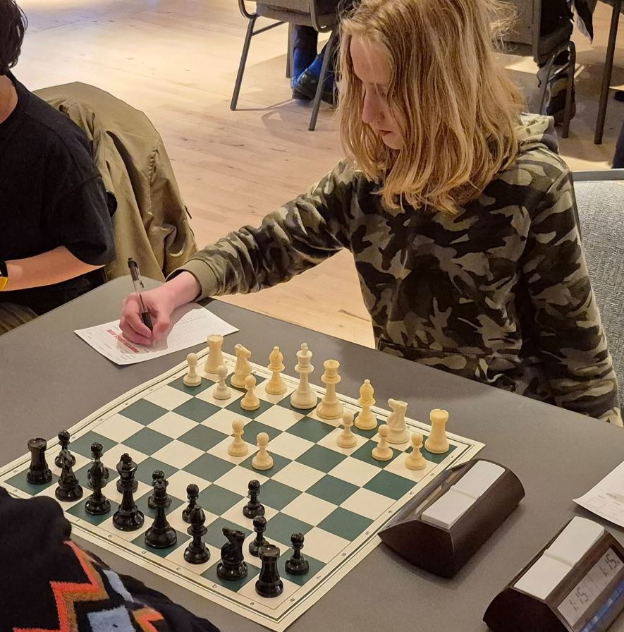 Junior player recording a move on a scoresheet beside a tournament chessboard and clock